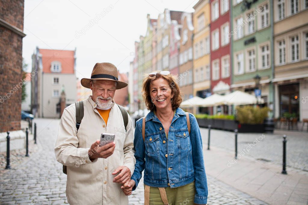 A portrait of happy senior couple tourists smiling, holding hands, using smartphone outdoors in historic town