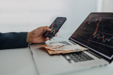 Bir işadamı ofis masasında bilgisayar ve akıllı telefonla çalışarak avro parası sayıyor, enflasyon kavramı.