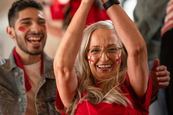 An excited senior woman football fan supproting English national team in live soccer match at stadium.