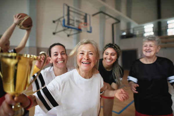 A group of young and old women, basketball team players, in gym with trophy celebrating victory.