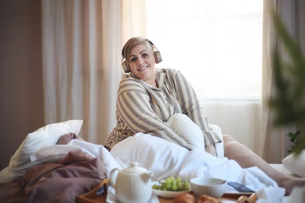 Glad Överviktig Kvinna Med Hörlurar Och Smartphone Äter Frukost Sängen — Stockfoto