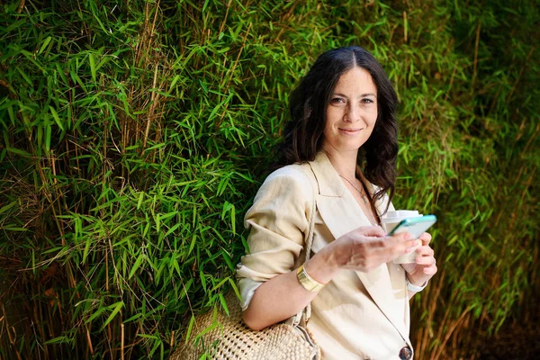 一位快乐的女性旅行者 一边用手机发短信 一边拿着咖啡杯 背景是绿色的植物 — 图库照片