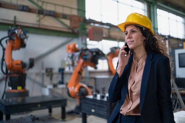 An automation female engineer with tablet for programming robotic arm calling on cellphone and walking through factory.