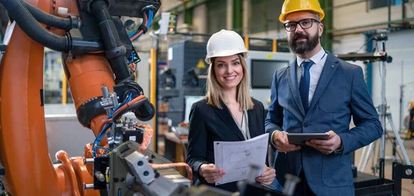 A chief engineer and project manager in modern industrial factory with robot arms talking and planning optimization of production.