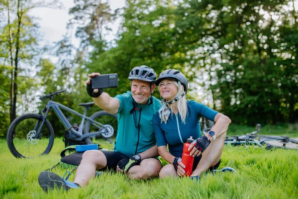 An active senior couple resting after bicycle ride at summer park, sitting on grass and taking slefie.