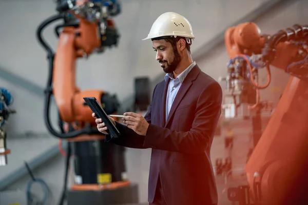 An automation engineer uses tablet for programming robotic arm in factory.