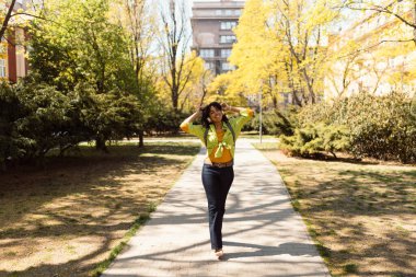Mutlu, gülümseyen, güzel, kıvırcık bir kadın parkta yürüyor kulaklıkla müzik dinliyor..