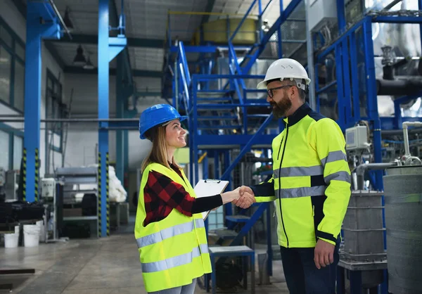 一位穿着制服的工程师和产业工人在大型金属工厂大厅里握手交谈 — 图库照片