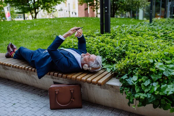 仕事中に都会の公園のベンチでリラックスしたヘッドフォンで音楽を楽しむ成熟したビジネスマン — ストック写真