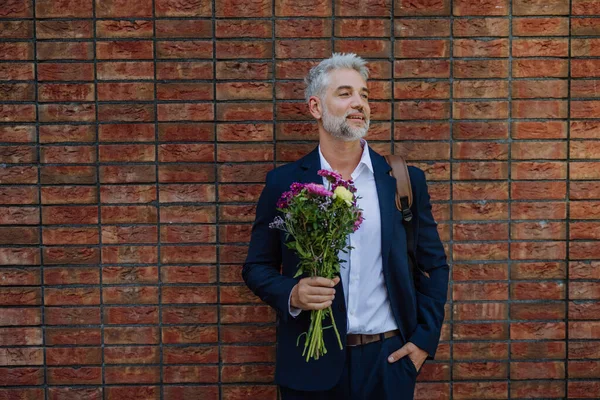 A portrait of happy mature man with flower bouquet standing in front of brick wall and waiting for girlfriend.