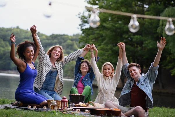 A group of young friends having fun on picnic near a lake, sitting on blanket and raising arms.