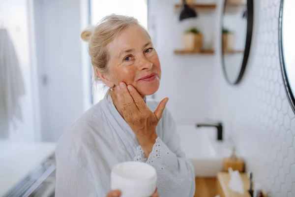 一位穿着浴衣的漂亮老年女士在浴室 皮肤护理和晨间日常活动中使用天然奶油 — 图库照片