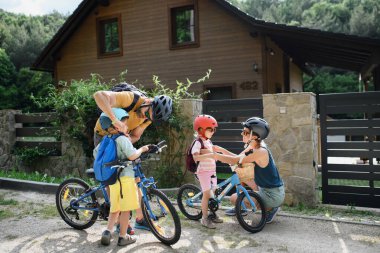Bisiklet sürmeye hazırlanan, evin önünde bisikletle duran küçük çocuklu genç bir ailenin portresi..