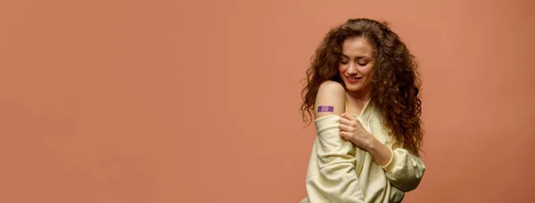 ワクチンを飲んだ女性の笑顔が描かれている 彼女のシャツの袖を保持し オレンジの背景に隔離されたワクチン接種を受けた後 包帯で彼女の腕を示し女性 — ストック写真