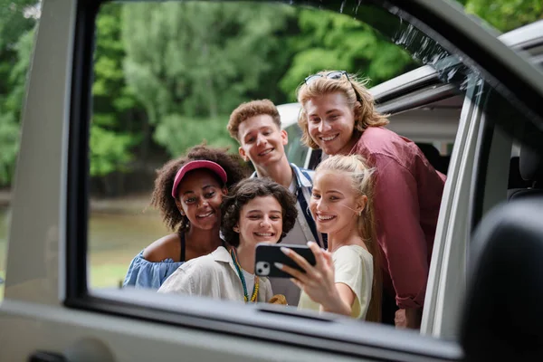 Çok ırklı genç arkadaşlar arabayla seyahat eder, selfie çekerler - yaz tatili, tatil, seyahat, yolculuk ve insanlar konsepti.