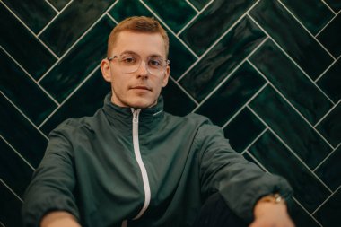 Ceketli ve gözlüklü genç bir adamın portresi. Kocaman, koyu renkli bir duvarda oturuyor..