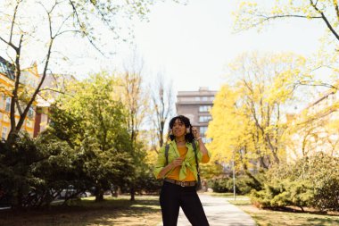 Mutlu, gülümseyen, güzel, kıvırcık bir kadın parkta yürüyor kulaklıkla müzik dinliyor..