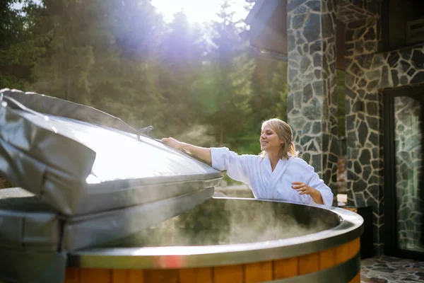 A woman in bathrobe opening lid of hot tub, checking temperature, ready for home spa procedure in hot tub outdoors. Wellness, body care, hygiene concept.