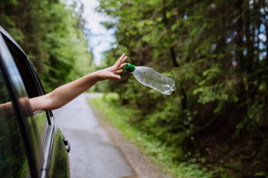 A womans hand throwing away plastic bottle from car window on the road in green nature, environmental protection, global warming concept