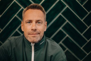 Ceketli orta yaşlı bir adamın portresi. Kocaman, koyu renkli bir duvarda oturuyor..