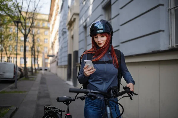 A portrait of businesswoman commuter on the way to work with bike, using smartphone, sustainable lifestyle concept.