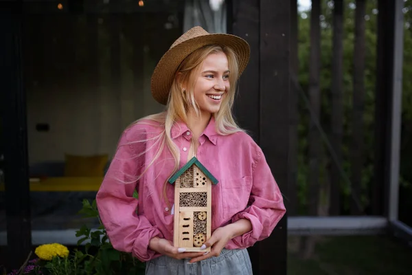 A young woman garedener holding bug and insect hotel on terrace in garden, sustainable lifestyle.
