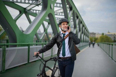 A businessman commuter on the way to work, pushing bike on bridge and calling on mobile phone, sustainable lifestyle concept.