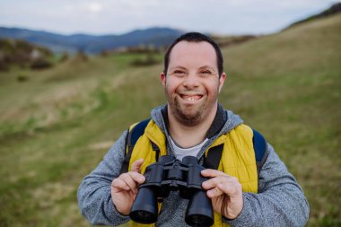 Portrait of happy young man with Down syndrome outdoor, enjoying nature and looking trough a binoculars.