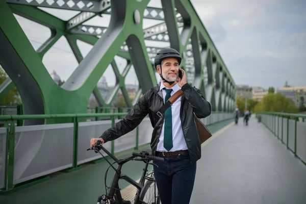 A businessman commuter on the way to work, pushing bike on bridge and calling on mobile phone, sustainable lifestyle concept.