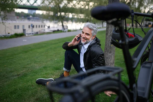 A businessman with bike sitting on grass in park, using smartphone. Commuting and alternative transport concept