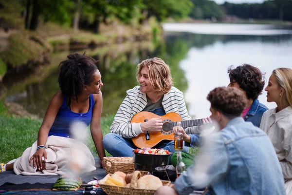 A group of young friends having fun on picnic near a lake, sitting on blanket eating and playing guitar.