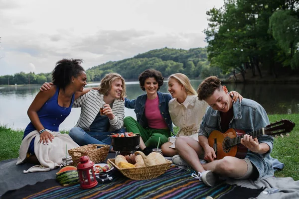A group of young friends having fun on picnic near a lake, sitting on blanket eating and playing guitar.