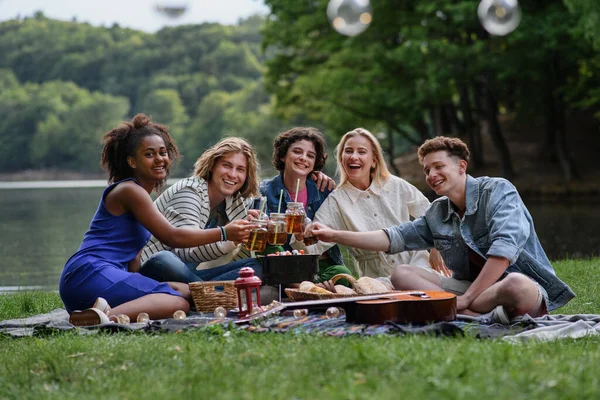 A group of young friends having fun on picnic near a lake, sitting on blanket and toasting with drinks. Looking at camera.