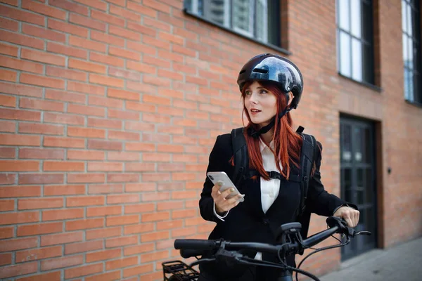A portrait of businesswoman commuter on the way to work with bike, using smartphone, sustainable lifestyle concept.