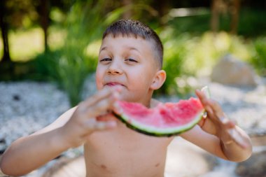 Little boy sitting near a lake and eating watermelon on hot sunny day during summer vacation.