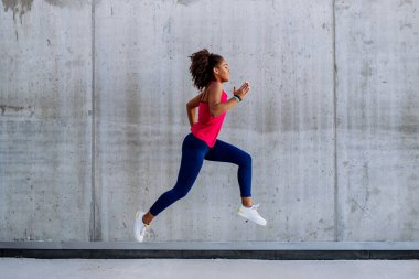 Young multiracial girl jogging and jumping in city, in front of a concrete wall, side view.