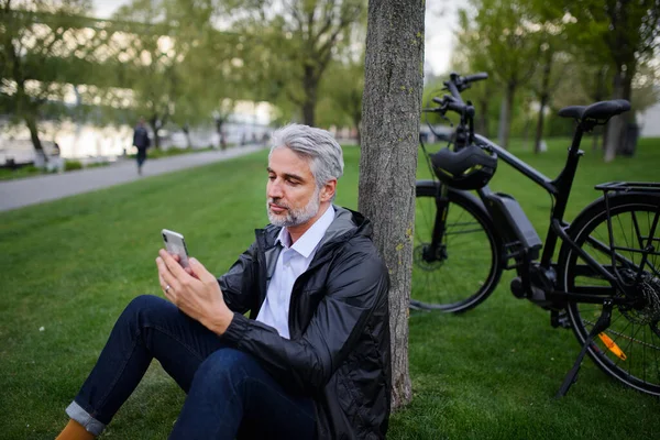 A businessman with bike sitting on grass in park, using smartphone. Commuting and alternative transport concept