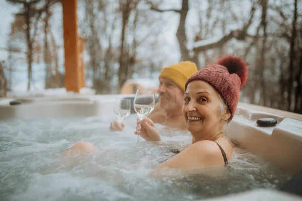 在寒冷的冬日里 头戴火帽的老夫妇在阳台上一起享受户外浴缸和酒杯的乐趣 — 图库照片