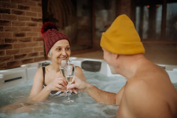 在寒冷的冬日里 头戴火帽的老夫妇在阳台上一起享受户外浴缸和碰杯的乐趣 — 图库照片