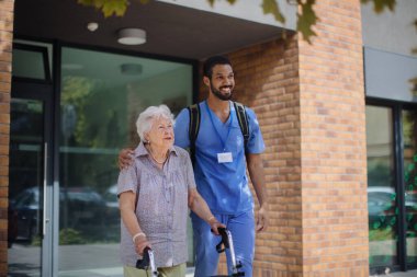 Caregiver walking with senior woman client in front of a nurishing home. clipart