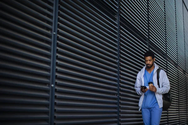 Young multiracial man working as nurse coming back from work, checking smartphone and holding cup of a coffee.