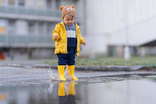 Happy little boy in yellow raincoat jumping in puddle after rain in cold autumn day.