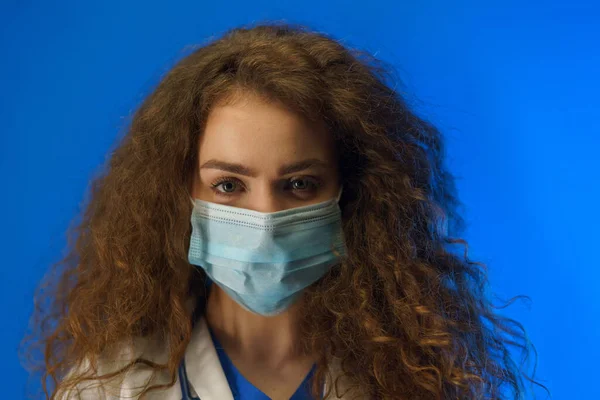 青い背景を背景に カメラを見て外科的顔のマスクを持つ若い女性医師の肖像画 — ストック写真