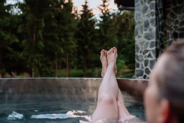 An unrecognizable young woman with feet up relaxing in hot tub outdoor in nature.