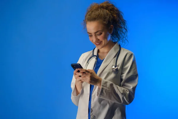 一个工作室用蓝色背景的手机拍摄的年轻女医生的照片 — 图库照片