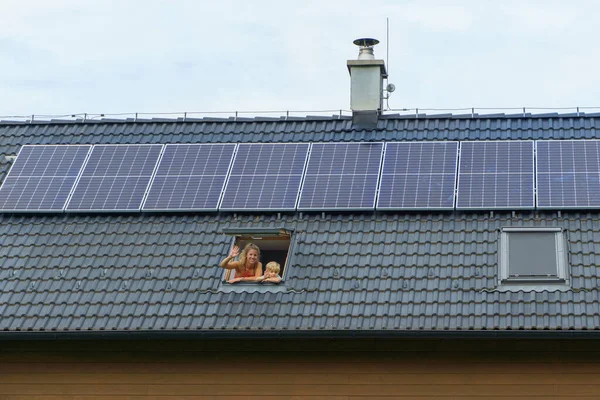屋根の上にソーラーパネルが設置された家の窓からは 幸せな母親と息子が手を振っています 代替エネルギー 省資源 持続可能なライフスタイルの概念 — ストック写真