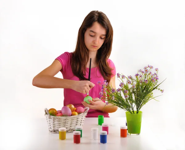 Girl painted Easter eggs. On the table is a basket with colored eggs, paint for coloring and a vase of flowers — Stock Photo, Image