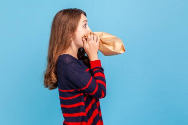 Gergin, stresli bir kadının çizgili, sıradan bir süveter giymesi, zar zor nefes alması, kese kağıdı kullanması, sakinleşmeye çalışması, kendini geliştirmesi. Mavi arka planda kapalı stüdyo çekimi.