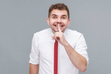 Bu bir sır. Beyaz gömlekli ve kravatlı genç bir adamın portresi. Parmakları dudaklarında, sırlarını ve gülümsemelerini söyle. Gri arkaplanda kapalı alan.