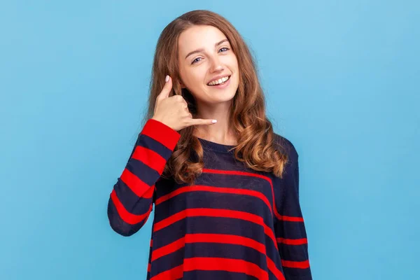 Hey handsome, call me. Smiling woman wearing striped casual style sweater flirting holding fingers near ear showing call gesture, answering call. Indoor studio shot isolated on blue background.